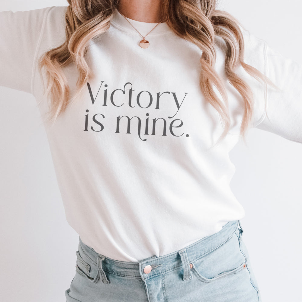 Blonde woman wearing a white Christian sweatshirt with "Victory is Mine" message in modern serif font, symbolizing faith and strength in a minimalist design